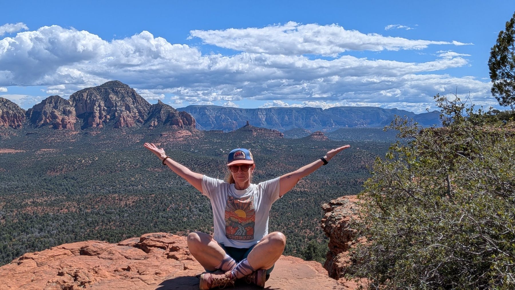 Marybeth sitting cross-legged with arms open wide on a red rock overlook in Sedona